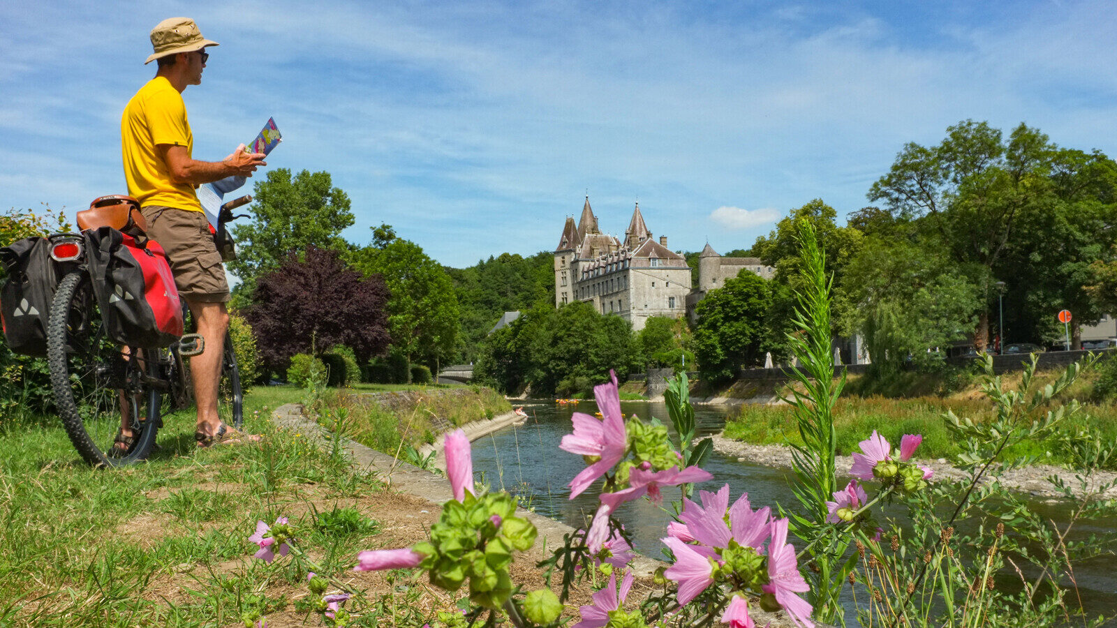 un homme se repose en consultant une carte adossé sur son vélo au bord d'une rivière, il observe un château en arrière plan